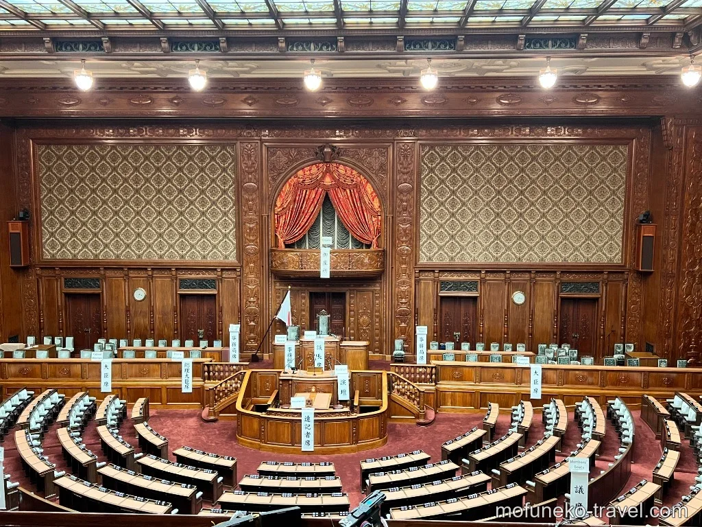 衆議院議場の全体写真。傍聴席から見下ろした議長席・演壇・各議員席の様子。正面の赤いカーテンのバルコニーが印象的。